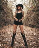 Woman in black cowboy hat, boots, and shorts standing in a forest path.