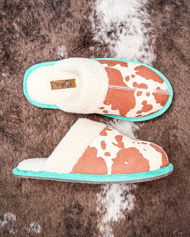 Pair of cow print slippers with turquoise trim on a fur surface