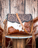 Brown and white cowhide handbag with floral tooled leather on a wooden stool against a wooden wall.