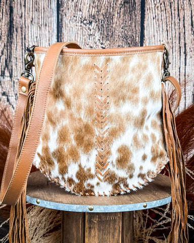 A brown and white hair-on-hide crossbody bag with a leather strap and fringe, displayed on a wooden surface.