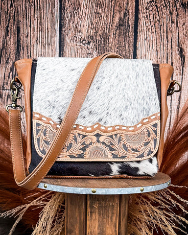 A brown and white cowhide crossbody bag with hand-tooled floral leather details and an adjustable strap, displayed on a wooden stool.