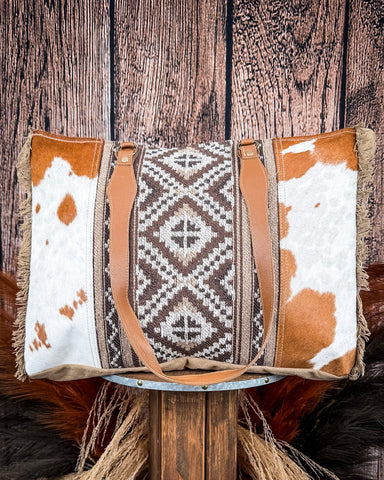 Cowhide-patterned bag with decorative patch on a wooden stand against a wooden background