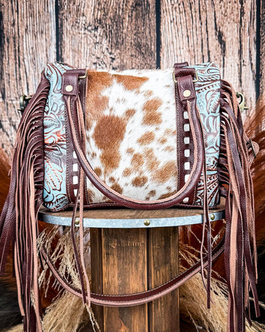 Cowhide leather handbag with fringe on a wooden stool against a rustic wooden background