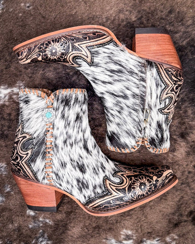 A pair of ankle booties with a snip toe design, featuring a mix of white and brown hair-on-hide leather, intricate floral tooling, and a stacked wooden heel.