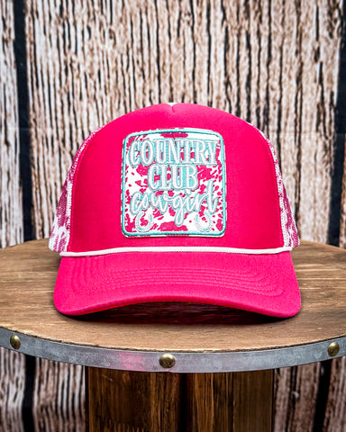 The Country Club Pink Cow Print Trucker Cap displayed on a wood table with a wood backdrop.