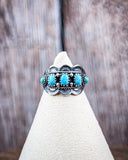 The Asia Adjustable Genuine Turquoise Ring on a display stand with a wood backdrop.