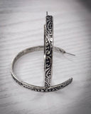 The Folk Tooled Hoop Fashion Earrings sitting on a wood table.