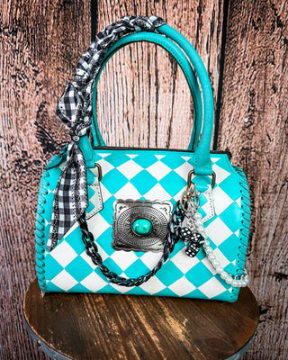 A turquoise and white checkered handbag with a black-and-white gingham scarf tied to the handle, a silver concho-style ornament, and a black-and-white checkered strap, displayed on a wooden stool against a rustic wood plank background.
