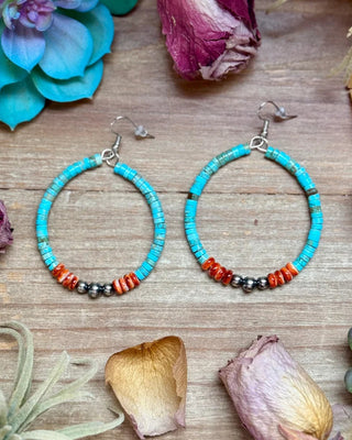 A pair of handmade hoop earrings with turquoise blue beads, small orange-red beads, and dark gray spacer beads, displayed on a rustic wooden surface with dried flowers and succulents.