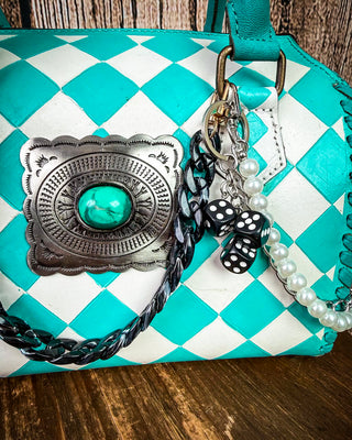 A turquoise and white checkered leather handbag with a large silver concho featuring a turquoise center, adorned with braided black and white strap, pearl beads, and a black-and-white polka dot bow charm on a wooden surface.