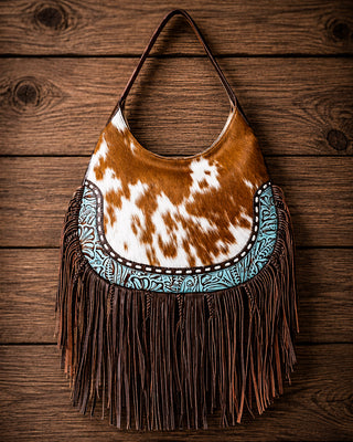 A western-style shoulder bag with a brown and white cowhide body, a turquoise and brown embroidered band, and long dark brown fringe, displayed against a rustic wooden plank background.