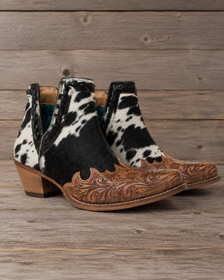A pair of women's western ankle boots featuring a black and white cowhide pattern on the upper and a hand-tooled brown leather vamp with floral scrollwork, set against a rustic wooden plank background.