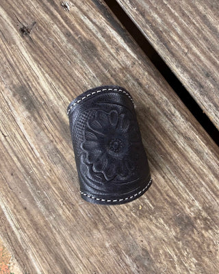 A black leather scarf slide with white contrast stitching and an embossed floral pattern, resting on a rustic wooden surface.
