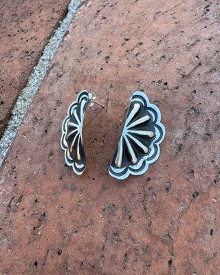 A pair of scalloped fan-shaped earrings with black and silver inlay, displayed on a textured reddish-brown stone surface.
