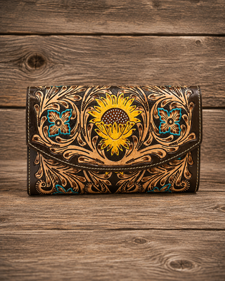 A tooled leather clutch wallet with a prominent yellow sunflower design and intricate brown floral scrollwork, accented with turquoise blue details, displayed on a rustic wooden surface.