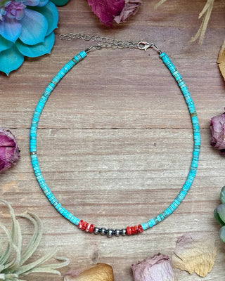 A beaded necklace with turquoise-blue beads, small red beads, and a cluster of dark gray beads, displayed on a wooden surface surrounded by dried flowers and succulents.