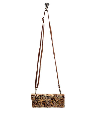 A brown leather crossbody bag with an intricate hand-tooled floral pattern, hanging from a black metal hook against a solid white background.