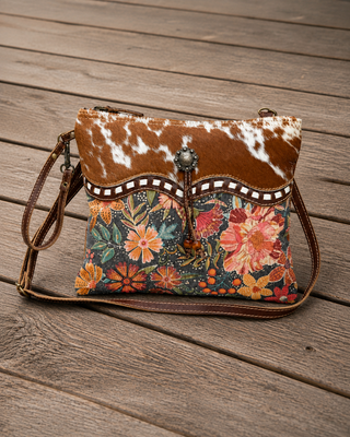 A western-style crossbody handbag with a brown and white cowhide flap, a decorative beaded trim, and a colorful floral fabric body, resting on a rustic wooden plank surface.