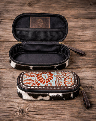Two zippered pencil cases with cowhide-patterned trim are displayed on a rustic wooden surface. The top case is open, showing a black interior and a brown leather patch with a western-style monogram logo.