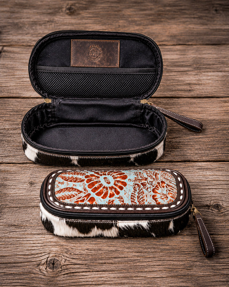 Two zippered pencil cases with cowhide-patterned trim are displayed on a rustic wooden surface. The top case is open, showing a black interior and a brown leather patch with a western-style monogram logo.