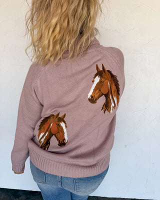 A woman viewed from behind wearing a dusty mauve knit sweater with embroidered brown horse heads on the back, paired with blue jeans against a white wall.