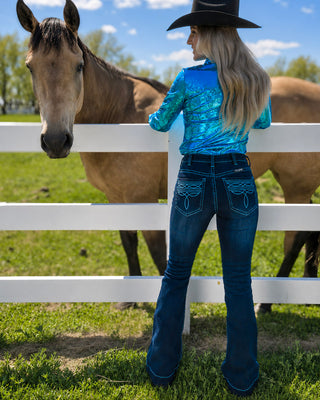 A woman in a black cowboy hat, a shiny blue long-sleeve top, and blue flared jeans stands behind a white fence in a grassy field, looking at a brown horse. The jeans feature decorative embroidered stitching on the back pockets.