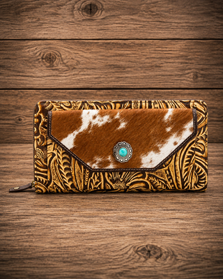 A western-style zip-around wallet with a tooled brown leather exterior and a cowhide flap featuring a turquoise concho accent, displayed on a rustic wooden background.