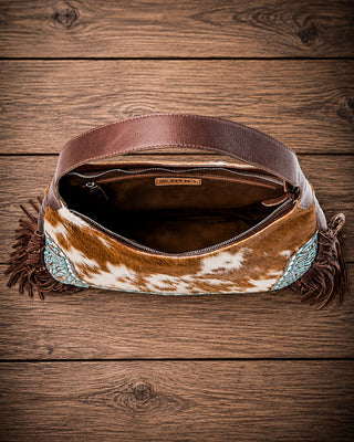 A western-style shoulder bag with a brown and white cowhide body, brown leather strap, dark brown fringe, and turquoise blue beaded accents, displayed open on a rustic wooden plank background.
