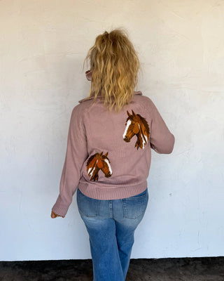 A woman viewed from behind wearing a dusty rose pink knit sweater with embroidered brown and white horse heads on the back, paired with blue jeans, standing against a white wall.