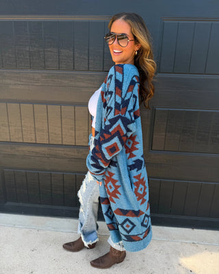 A woman wearing a blue knit cardigan with a red and dark gray geometric pattern, standing in front of a dark garage door.