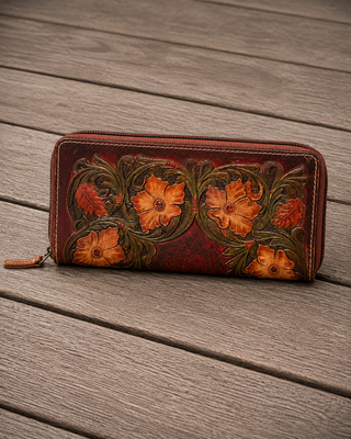 A brown leather zip-around wallet with hand-tooled floral designs in orange and green, resting on a gray wooden plank surface.