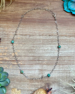 A delicate silver chain necklace with five small green beads spaced along the chain, laid flat on a rustic wooden surface surrounded by decorative succulents and dried flowers.