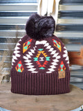 The Aztec Pom Beanie displayed on a wooden table with wheat in the back in front of a metal wall.