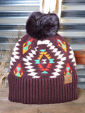 The Aztec Pom Beanie displayed on a wooden table with wheat in the background in front of a metal wall.