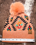 The Aztec Pom Beanie, shown on a cowhide backdrop with a ruler indicating it measures about 9 inches across.