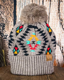 The Aztec Pom Beanie displayed on a wooden table with a wood background.
