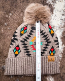 The Aztec Pom Beanie, shown on a cowhide backdrop with a ruler indicating it measures about 9 inches tall.
