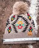 The Aztec Pom Beanie, shown on a cowhide backdrop with a ruler indicating it measures about 9 inches across.