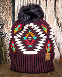 The Aztec Pom Beanie displayed on a wooden table with a wood background.
