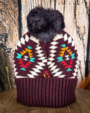 The Aztec Pom Beanie displayed on a wooden table with a wood background.