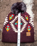 The Aztec Pom Beanie, shown on a cowhide backdrop with a ruler indicating it measures about 9 inches tall.