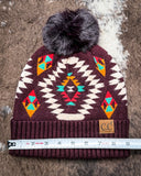 The Aztec Pom Beanie, shown on a cowhide backdrop with a ruler indicating it measures about 9 inches across.