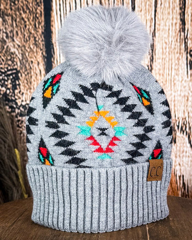 The Aztec Pom Beanie displayed on a wooden table with a wood background.