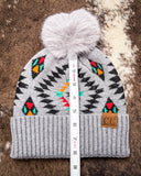 The Aztec Pom Beanie, shown on a cowhide backdrop with a ruler indicating it measures about 8 inches tall.