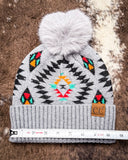 The Aztec Pom Beanie, shown on a cowhide backdrop with a ruler indicating it measures about 9 inches across.