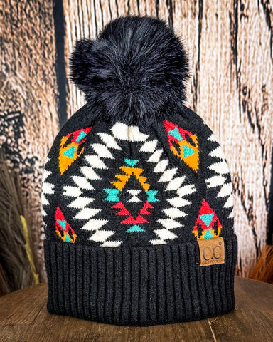 The Aztec Pom Beanie displayed on a wooden table with a wood background.
