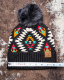 The Aztec Pom Beanie, shown on a cowhide backdrop with a ruler indicating it measures about 8 inches across.