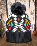 The Aztec Pom Beanie displayed on a wooden table with a wood background.
