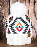 The  Aztec Pom Beanie displayed on a wooden table with a wood backdrop.