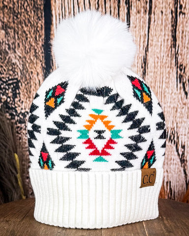 The  Aztec Pom Beanie displayed on a wooden table with a wood backdrop.